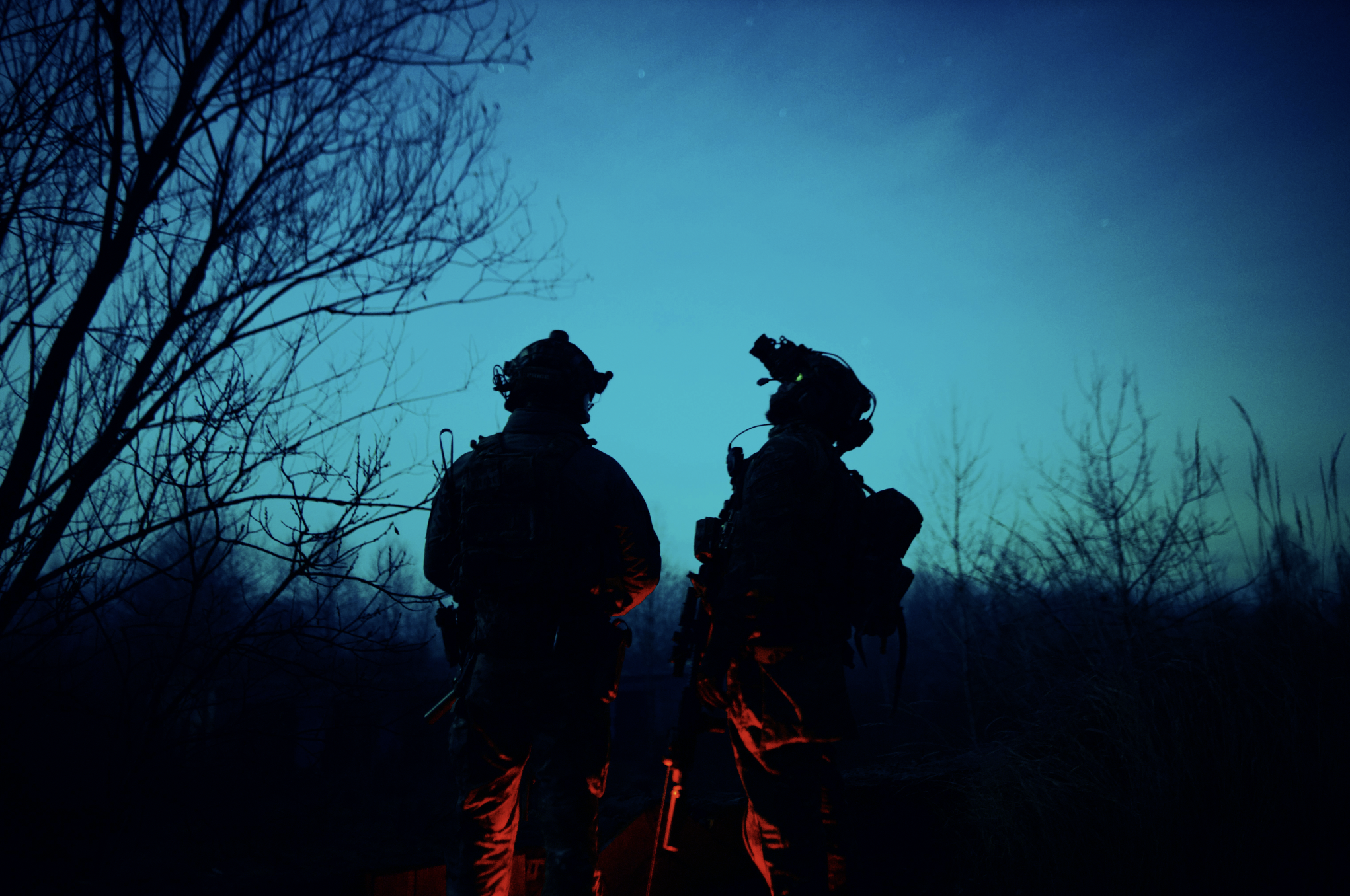 Nighttime scene of two warfighters in silhouette staring up at a deep blue sky.